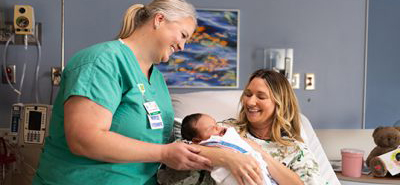 A nurse smiles while checking on a female patient and a newborn baby in a hospital bed. Family Birth Place