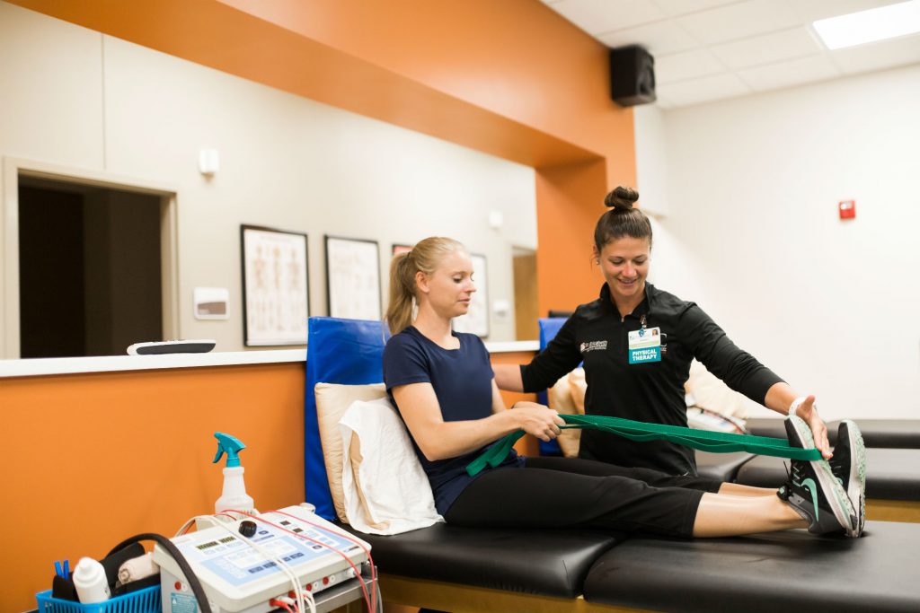 Blonde female patient stretches Achilles tendon using resistance band with the help of physical therapist.